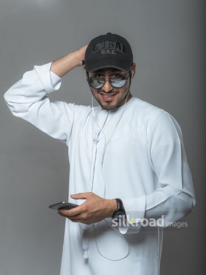 Arab man wearing a cap dressed in the traditional attire using his mobile|-
