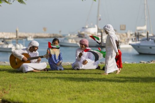 Arab boy walking around the group of people who are playing the guitar and singing along for national day|-
